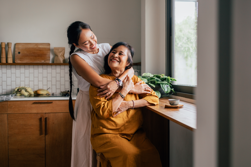 A Happy Beautiful Woman Hugging Her Mother While She Is Sitting In The Kitchen And Drinking Tea - stock photo
A smiling Asian woman enjoying drinking tea while her loving daughter is embracing her.
